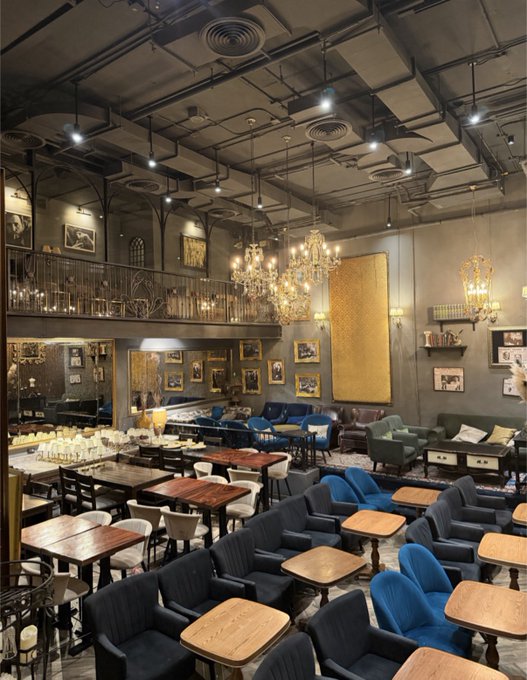 An empty, vintage-style cafe or bar interior with dark walls, ornate chandeliers, blue velvet chairs, and wooden tables.