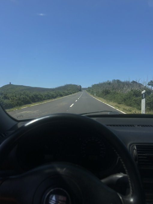 A driver's view from behind the steering wheel of a Volkswagen (logo visible on the wheel) traveling down an empty rural road under a clear blue sky, seemingly heading off into the distance—perhaps toward a new destination or opportunity. The open road serves as a metaphor for the call to "drop your SOL address," suggesting readiness to embark on a fresh journey.