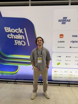 A man in a gray shirt and khaki pants smiles as he poses in front of a "Block|chain.RIO" backdrop featuring sponsor logos including SEBRAE, Banco Original, Visa, Goote, Z.ro Bank, and Mercado Bitcoin. The event signage and his excited presence reflect the post's enthusiasm for the community advancing currency competition through blockchain innovation.