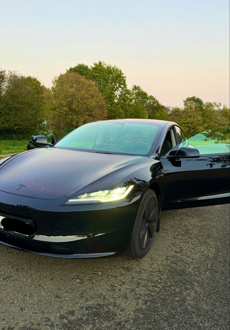 A sleek black Tesla car parked outdoors with its driver's side door open, showcasing its illuminated headlights against a backdrop of trees at sunset.