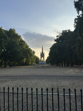 A scenic view looking through a black metal fence towards a large stone monument, framed by large green trees under a partly cloudy sky.