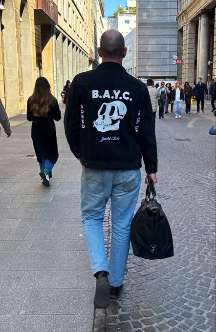 A street-level photo of a man walking away on a cobblestone street, wearing a black Bored Ape Yacht Club (BAYC) denim jacket, light blue jeans, and carrying a black duffel bag.