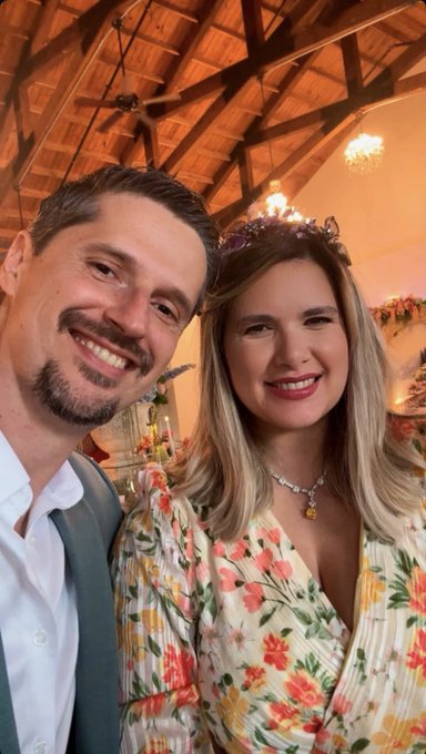 A selfie of a smiling couple at what appears to be a wedding or formal celebration; the man has dark hair and a beard, and the woman wears a floral dress with a floral crown, in a venue with a wooden ceiling and chandeliers.