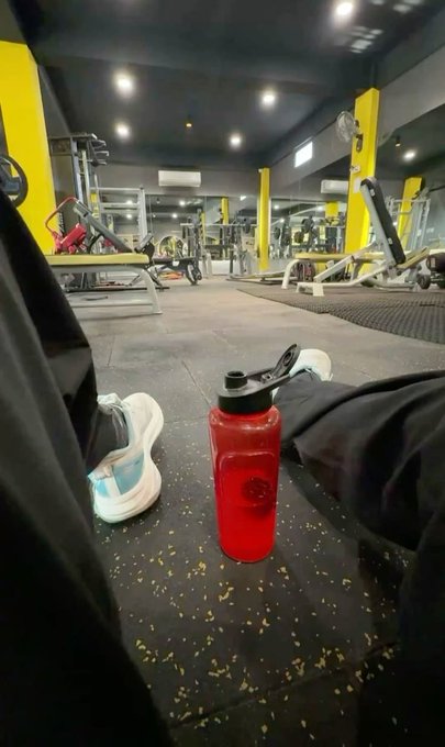 A person sits on a gym floor with a red water bottle nearby, showing gym equipment in the background.