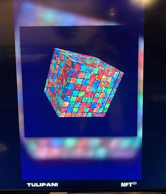 A digital screen displays a colorful 3D cube NFT artwork composed of multicolored textured squares against a dark blue background, with 'TULIPANI' and 'NFT' logos at the bottom.