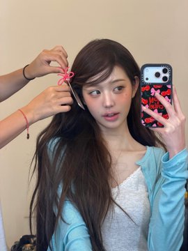 The image shows a young woman taking a mirror selfie with a red heart-patterned phone case while someone behind her ties a pink ribbon bow into her long brown hair, embodying the soft and playful "mintcore" aesthetic referenced in the caption.