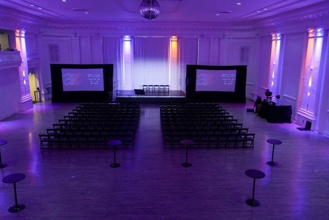 A purple-lit event hall with rows of black chairs set up before a stage with two projection screens and a small panel table.