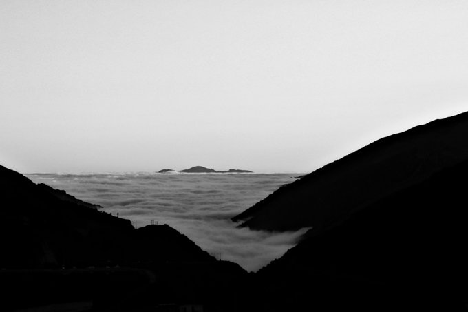 The image shows a beautiful landscape with mountains and a valley below, creating an impression of a serene and peaceful environment. The sky above appears to be cloudy, adding depth to the scene. In the background, there are islands visible through the clouds, suggesting that this location is near a body of water or a coastal area. The image also contains text elements such as "Morning friends Happy Sunday Before the world woke, an island lay in wait for the sun." and "@objktcom". These details provide context to the scene and help convey the overall atmosphere of the photograph.