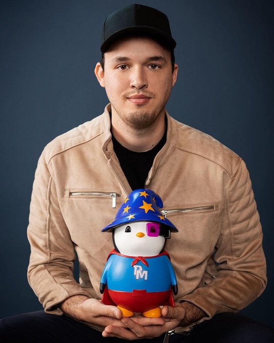 A man wearing a black cap and tan jacket holds a small penguin toy dressed in a wizard hat, purple eye patch, and a red and blue outfit with an 'RM' logo, against a dark blue background.