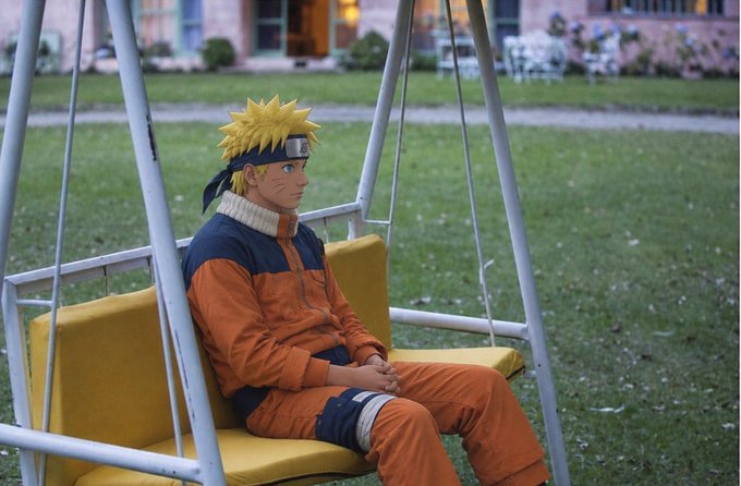 A person in a Naruto Uzumaki cosplay, complete with spiky blonde wig and orange jumpsuit, sits on a yellow outdoor swing.