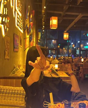 The image features a woman sitting at a table with her hand making a peace sign, possibly as part of a selfie or social media post. The background includes a restaurant setting with hanging lamps and other people present. There are also some food items on the table, such as bowls and cups. The overall atmosphere suggests that this is an indoor dining area where people gather to enjoy meals and spend time together.