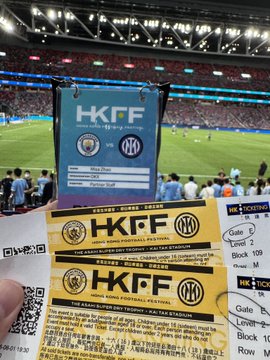 A hand holds two yellow 'HKFF - Hong Kong Football Festival' tickets for a Singapore vs. Inter Milan match, with seat details 'Gate F Level 2 Block 109 Row M Seat 13' visible in a stadium setting.