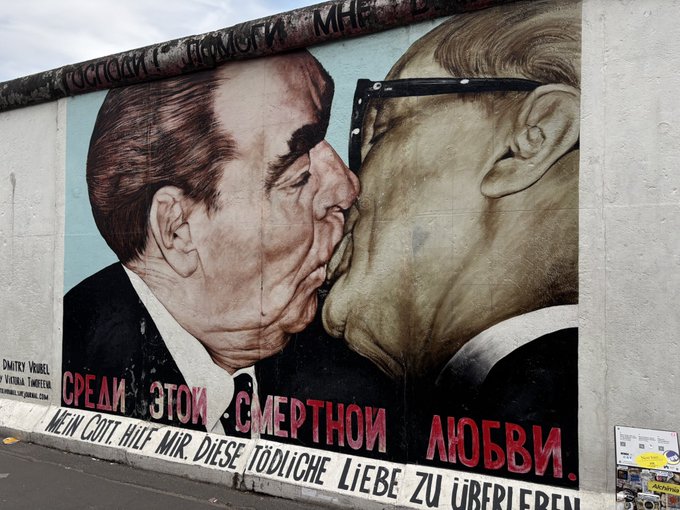 The image features a large mural painted on a wall that depicts two men kissing. The mural is located next to a street where there are cars parked, indicating an urban setting. The caption of the image reads "Focus on what is in the image and how it relates to the post".