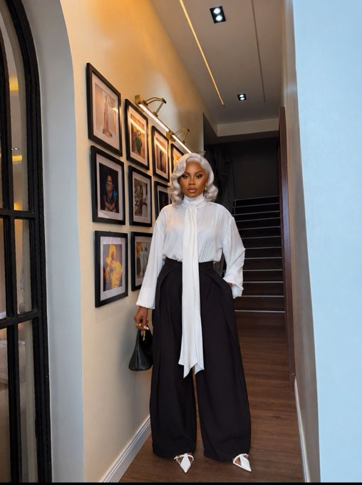A stylish woman with silver hair posing in an elegant hallway, wearing a white tie-neck blouse and high-waisted black wide-leg trousers with white pointed heels.