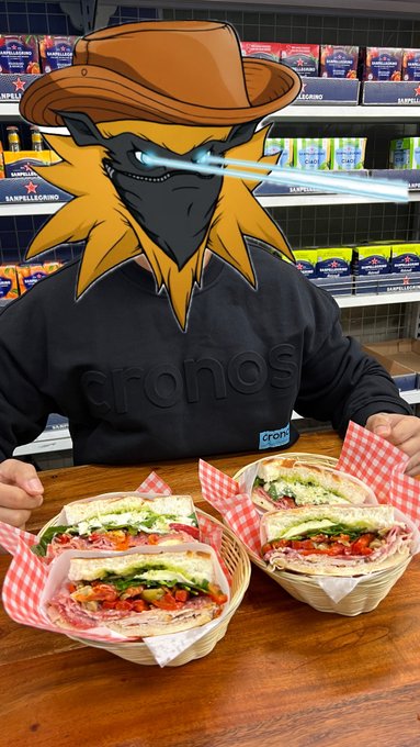 A person in a 'cronos' sweatshirt with a cartoon character filter featuring a yellow creature in a cowboy hat with laser eyes covering their face, sitting at a table with four baskets of sandwiches in front of shelves stocked with Sapporo beer and other products.