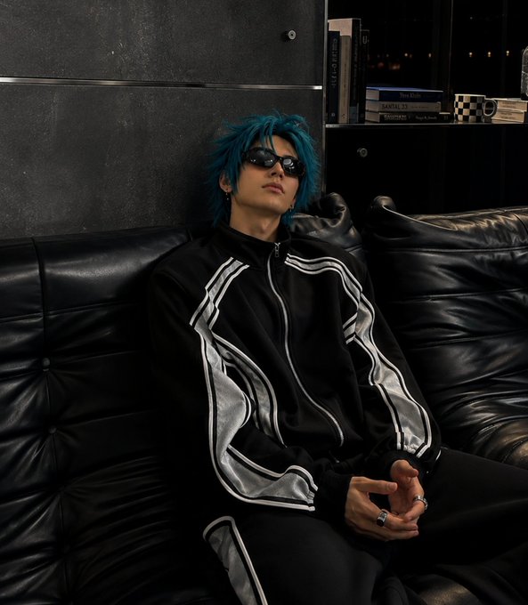 A young person with teal blue hair and sunglasses, wearing a black and white track jacket, reclining on a black leather couch with bookshelves visible in the dimly lit background.