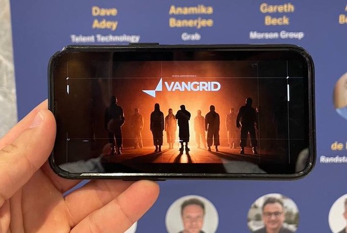A hand holds a smartphone displaying the 'VANGRID' logo against a backdrop of silhouetted figures, with a background poster listing names like Dave Adey, Anamika Banerjee, and Gareth Beck.