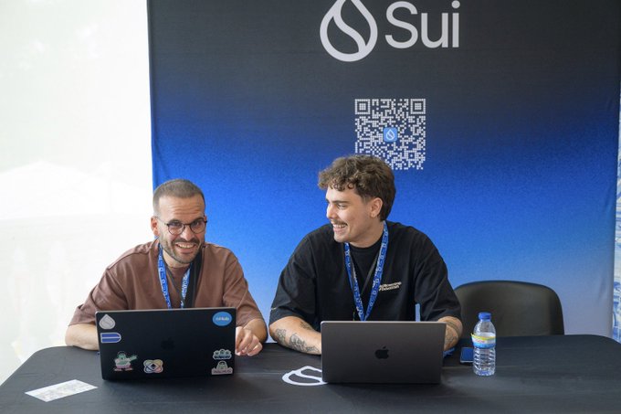 Two men sit at a table smiling and working on laptops, with a blue 'Sui' backdrop featuring a QR code behind them.
