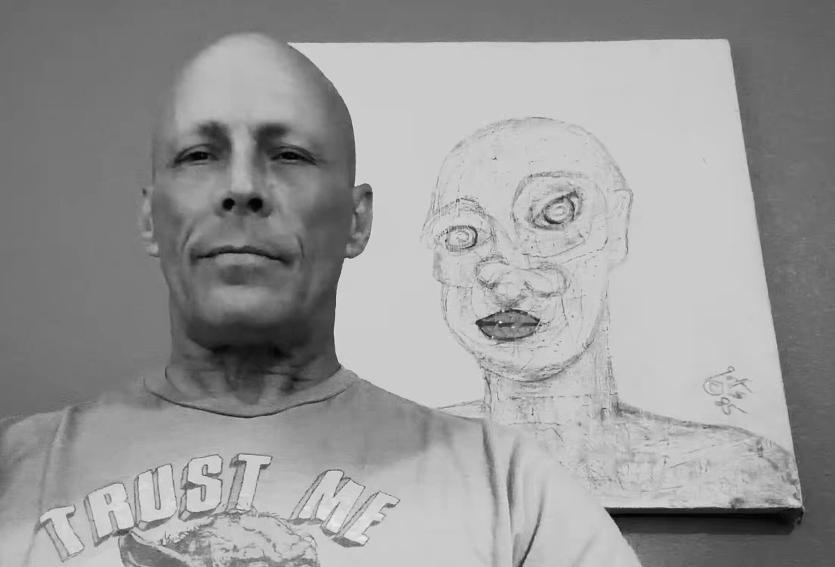 The image features a man wearing a gray t-shirt with "Just Me" written across the front, standing next to an art piece that depicts a face of a woman. The man appears to be posing for the photo while looking at the artwork. The background is a simple gray wall, and there are no other objects or people in the scene.