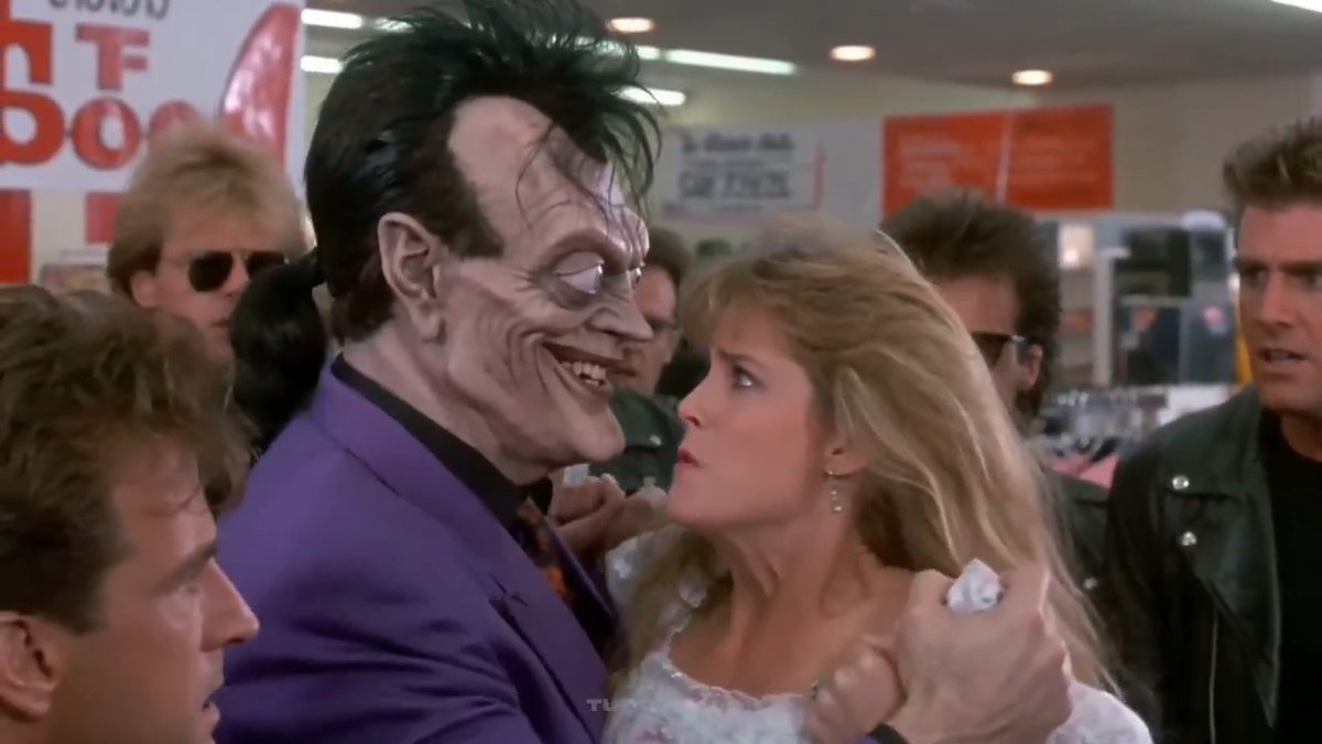A sinister clown-like villain with a grotesque grin in a purple suit grabs a frightened blonde woman in a white dress by the shoulders, surrounded by onlookers in what appears to be a store setting. This iconic scene from the Batman film (1989), featuring the Joker menacingly confronting his hostage, serves as a visual metaphor for the unpredictable and potentially threatening reality of memecoins—suggesting that what seems like fantasy investment can quickly materialize into a real, looming danger.