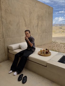 A young man sits casually on a concrete ledge in a minimalist desert setting, holding a drink, with a tray of food and a pair of sandals beside him. The serene, solitary scene captures the "aurafarming" vibe mentioned in the post—suggesting a quiet moment of leisure away from his $300 million crypto gains.