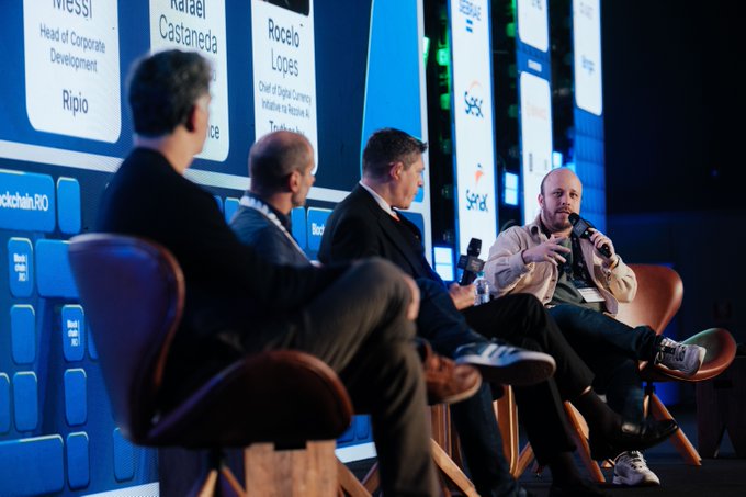 A panel discussion on stage featuring four men seated in chairs, with large screens in the background displaying names and titles including 'Rafael Castaneda', 'Rocelo Lopes', and 'Ripio'.