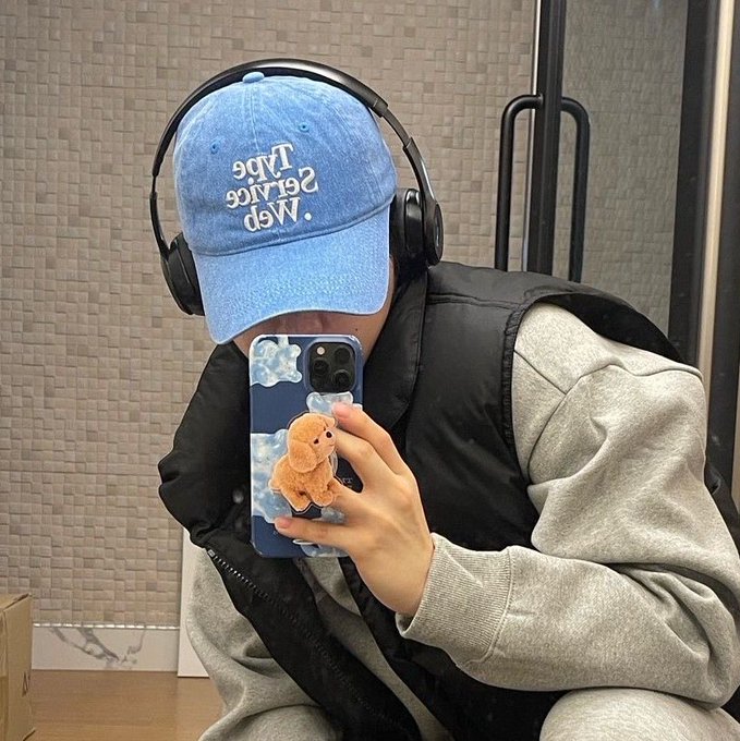 A person wearing a blue baseball cap with "Map space table" text, headphones, and a vest takes a mirror selfie with an iPhone that has a teddy bear case.