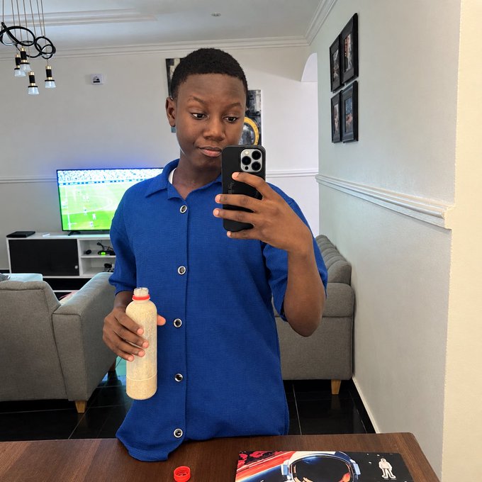 A young man in a blue button-up shirt takes a mirror selfie with a smartphone in a modern living room, holding a bottle of what appears to be a milky beverage, with a TV showing a football match and framed photos on the wall behind him.