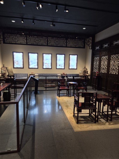 The interior of a traditional Chinese room or museum display featuring dark wood furniture, chairs, and decorative lattice work, with several framed pictures hanging on the back wall.