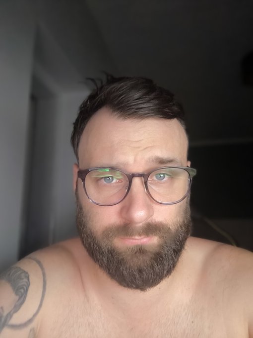The image features a man wearing glasses with a beard who has decided to take a selfie while standing naked against a gray backdrop. His shirtless appearance, along with his glasses and beard, sets him apart from the typical selfie-taker as he poses for the camera in an unconventional manner.