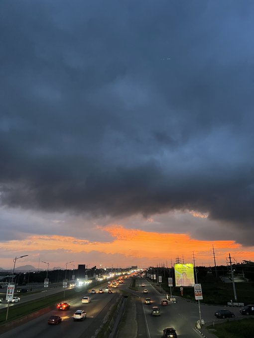 A dramatic sunset over a busy multi-lane highway with dark storm clouds above and vibrant orange light on the horizon, with car headlights visible on the road.