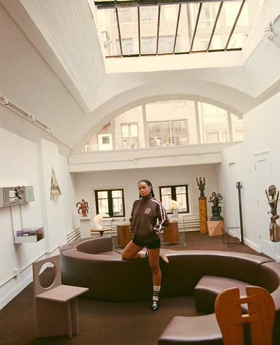 A fashion editorial photograph of a young woman in a brown oversized sweatshirt and black shorts standing inside a spacious, white-walled loft featuring a skylight, arched window, and a curved brown sofa.
