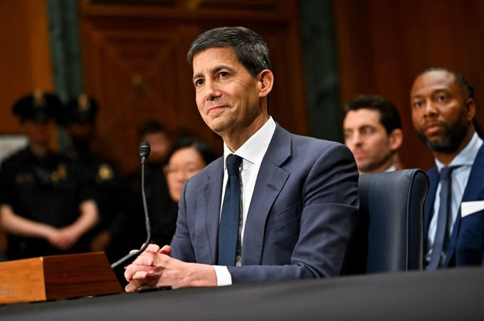 A man in a navy suit sits with his hands clasped at a formal hearing, with a uniformed officer and other officials visible in the background. The image appears to depict a government or financial official, though no connection to the CPI inflation data or Bitcoin price discussion in the caption is evident.