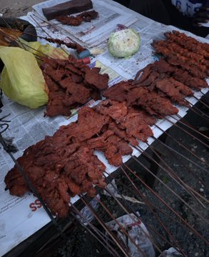 The image displays an outdoor street food stall with skewered, deep-red grilled meat (likely suya or kilishi-style spiced beef) laid out on newspaper, alongside an onion and yellow wrapper, with partial text "S S" visible on the paper. The caption ties into the post by humorously showing someone spending their last money on street food despite the cold weather, referencing slang like "Suga" and the memecoin tickers $ANSEM and "African Bull" as financial alternatives.