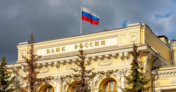 The Bank of Russia (Банк России) headquarters building with the Russian tricolor flag flying above it under a partly cloudy sky, surrounded by evergreen trees.