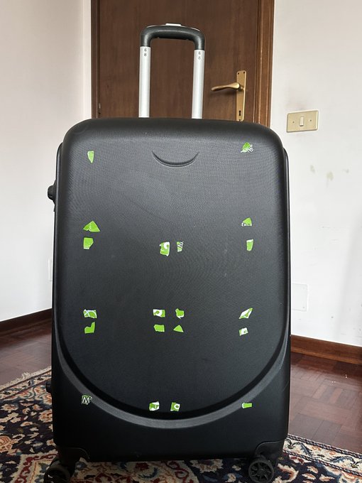 A black hard-shell suitcase stands on a wooden floor, decorated with scattered green and white stickers arranged in a smiley face pattern.