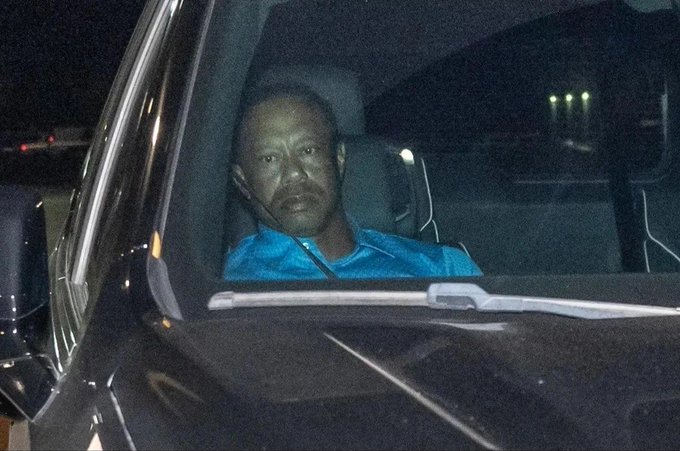 A man in a blue shirt sits inside a vehicle at night, visible through the window with a serious expression.