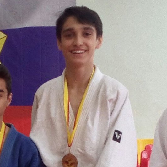 A smiling young man wearing a white martial arts gi with a yellow-trimmed collar and a bronze medal around his neck stands in front of a flag.