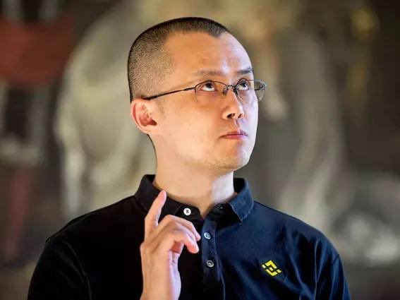 A man wearing glasses and a black polo shirt with a Binance logo, gesturing with his hand in what appears to be an interview setting.