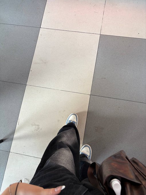 A first-person view looking down at someone's feet wearing white and blue sneakers, dark jeans, and carrying a brown leather bag while standing on gray tiled flooring.