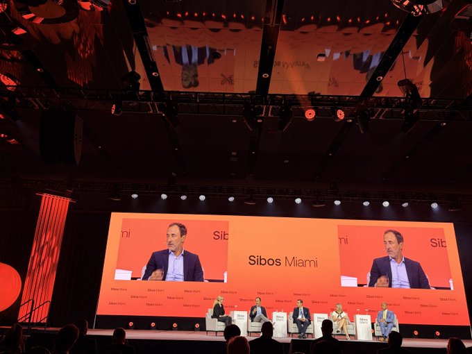 ENS team members sit on a panel at Sibos Miami, where a large screen displays the event branding alongside a speaker in a blue suit. The scene captures a financial conference focused on digital identity and onchain infrastructure.