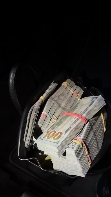 A photograph of a seat pocket on an airplane or train filled with thick stacks of $100 bills bound together with colorful rubber bands, set against a dark background.