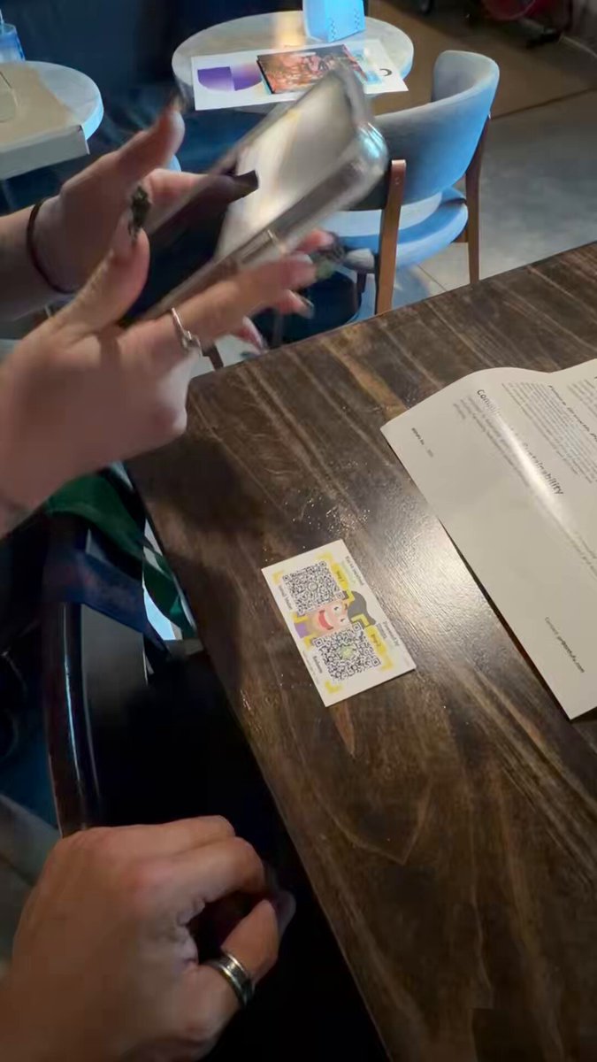 In this image, two people are sitting at a wooden table with a cell phone open and being used as a tool for demonstrating Bitcoin Cash. One person is holding up the phone while another person looks on, possibly learning about or considering adopting the technology. The scene suggests that they might be discussing the process of adopting Bitcoin Cash, which could involve using their phones to show others how it works or to demonstrate its benefits and features.