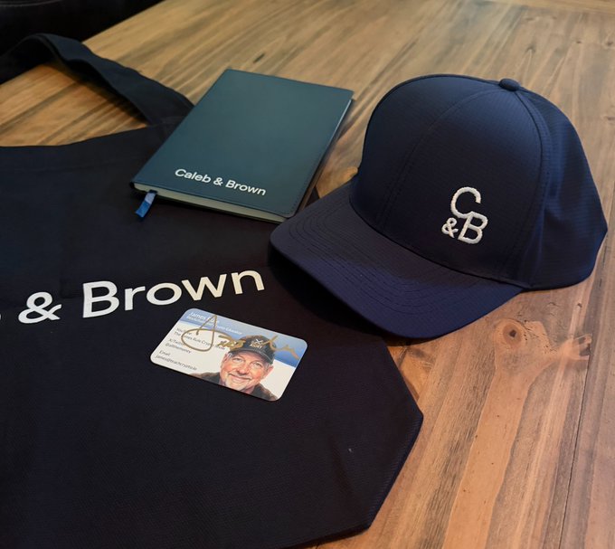 A black tote bag, baseball cap, and notebook branded with the "Caleb & Brown" logo sit on a wooden table alongside an ID card featuring a man's photo. The branded merchandise relates to the promotional giveaway mentioned in the post, where Caleb & Brown is offering prizes to four winners.