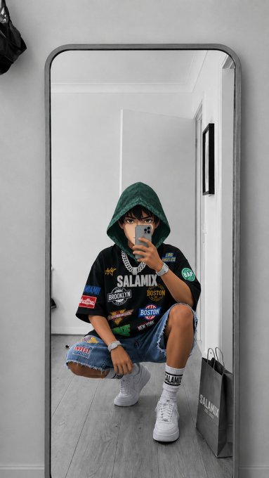 A person taking a mirror selfie while crouching, wearing a green hoodie, a black graphic t-shirt with 'SALAMIX', 'BROOKLYN', and 'BOSTON' patches, denim shorts, and white sneakers, holding a phone.