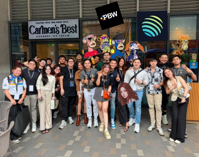 The image shows a group of people posing for a photo outside of a store, possibly an ice cream shop or a clothing store. The group consists of at least 13 individuals standing close together, with some holding up signs that say "PBW" and "PBW". There is also a person wearing a fake face on the right side of the image. A handbag can be seen near one of the people in the middle of the group. The store appears to have a window display featuring a cartoon character or a mascot, which adds an interesting visual element to the scene.