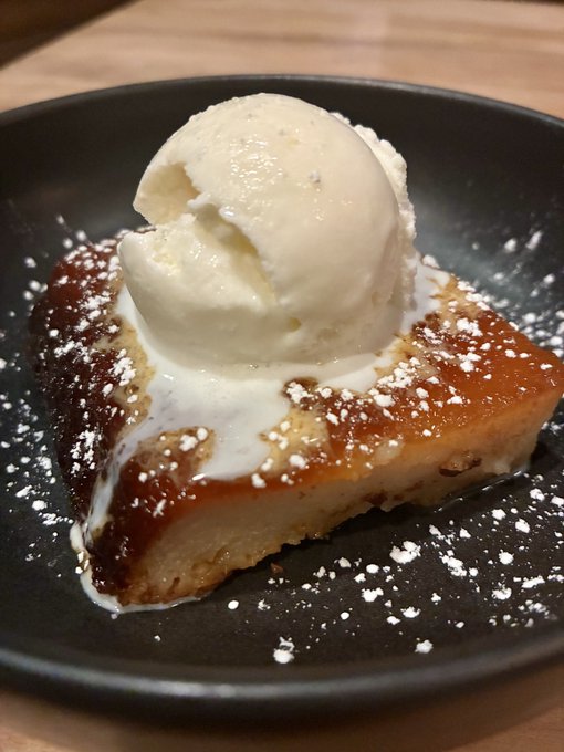 A slice of caramel-topped bread pudding or cake served on a black plate, topped with a scoop of vanilla ice cream and dusted with powdered sugar.