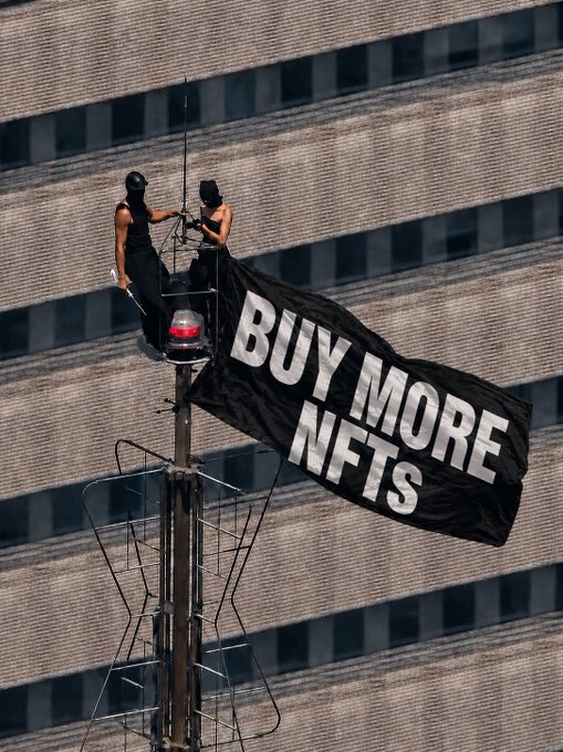 Two masked figures in black attire perch atop a transmission tower, unfurling a large black banner reading "BUY MORE NFTS" against the backdrop of a windowed skyscraper, embodying the crypto/NFT advocacy spirit associated with the Bored Ape Yacht Club community.