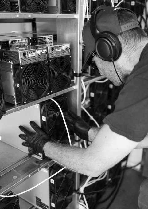 A black and white photo of a man wearing headphones and gloves, working on a rack of computer hardware with cooling fans, likely involved in cryptocurrency mining.