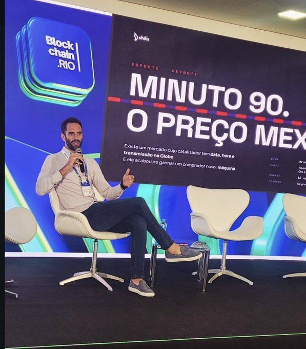 A man in a light shirt and jeans speaks on stage at the Blockchain Rio event, seated next to a large screen displaying a keynote titled 'MINUTO 90. O PREÇO MEX' (chiliz) about tokenization on the Globo network.